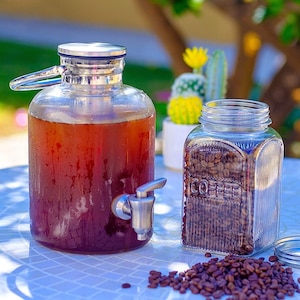 May include: A glass dispenser with a silver lid and spout filled with iced coffee. A glass jar filled with coffee beans sits next to the dispenser on a white tiled surface.  A small pile of coffee beans is scattered on the surface.