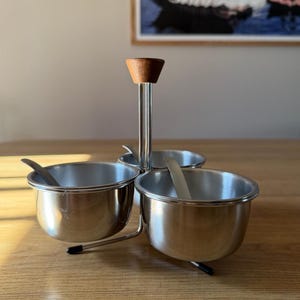 May include: A stainless steel condiment set with three bowls and small spoons. The set has a central handle with a wooden top. The bowls are arranged in a circular pattern, perfect for serving sauces or spices.