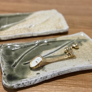 May include: A gold wristwatch with a white oval face and a mesh band rests on a rectangular ceramic dish. The dish has a speckled, off-white border and a green and beige interior. Another similar dish is in the background.