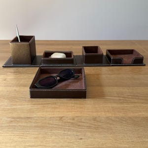 May include: A brown leather desk organizer set with white stitching. The set includes a pen holder, small trays, and a rectangular tray. A pair of black sunglasses rests in one of the trays. The organizer is on a wooden surface.