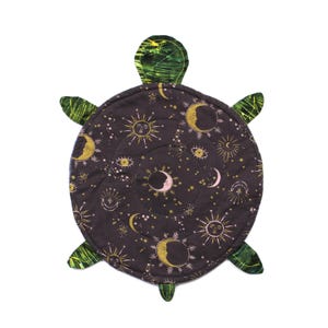 May include: A turtle-shaped fabric mat featuring a dark purple background with a gold and white celestial print of suns, moons, stars, and eyes. The head and four flippers are made from a green and black marbled fabric with visible stitching.