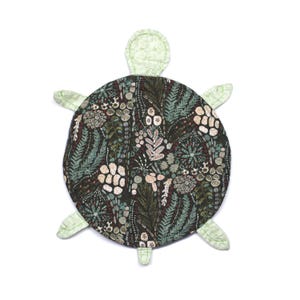 May include: A fabric turtle-shaped mat featuring a dark brown shell patterned with botanical illustrations of ferns, leaves, and circular shapes in shades of cream, sage green, and black. The head and four flippers are a light green fabric.