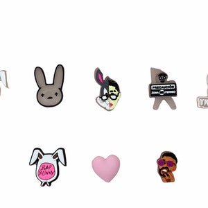 May include: Set of eight colorful cartoon character shoe charms featuring a pink heart, a cartoon man with sunglasses, a white rabbit with the text "Bad Bunny", a gray rabbit with a black X on its face, a gray rabbit with the text "YHLQMDLG", a gray rabbit with the text "Bad Bunny" on a pink balloon, a gray rabbit with a black and white Bugs Bunny face, and a gray stick figure with the text "Precaucion Zona Perro".