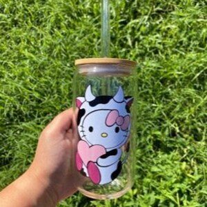 Peut inclure: Un gobelet en verre transparent avec un motif de vache noir et blanc repr&eacute;sentant Hello Kitty tenant un c&oelig;ur rose. Le gobelet est dot&eacute; d'un couvercle en bambou et d'une paille en plastique transparente.