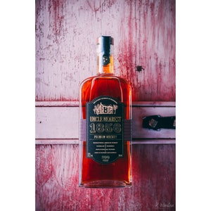 Puede incluir: Una botella de vidrio transparente de whisky premium Uncle Nearest 1856. La botella tiene una etiqueta negra con letras doradas y una pequeña ilustración de una casa. La botella está sobre una superficie de madera rosa.