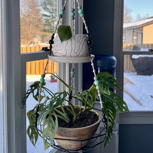 May include: A macrame plant hanger with black and white beads and light green beads hangs from the ceiling in front of a window. Two potted plants hang from the hanger. The top plant is in a white ceramic pot with a brown swirl design. The bottom plant is in a brown wooden pot and is a monstera plant.