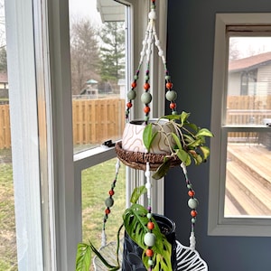 May include: A two-tiered plant hanger made of white beads with green and orange accents. The hanger is holding two potted plants, one with a white and brown pot and the other with a black pot. The hanger is hanging from a window.