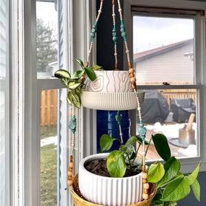 May include: Two white ceramic hanging planters with green plants, suspended from a macrame-style beaded cord with brown and turquoise beads. The planters are hanging in front of a window with a view of a snowy backyard.