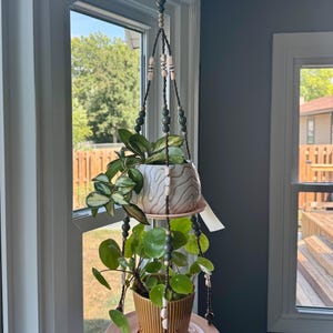 May include: A hanging plant holder with two ceramic pots and a pink plate. The holder is suspended from a hook and adorned with beads. The pots contain green plants, creating a decorative indoor display.