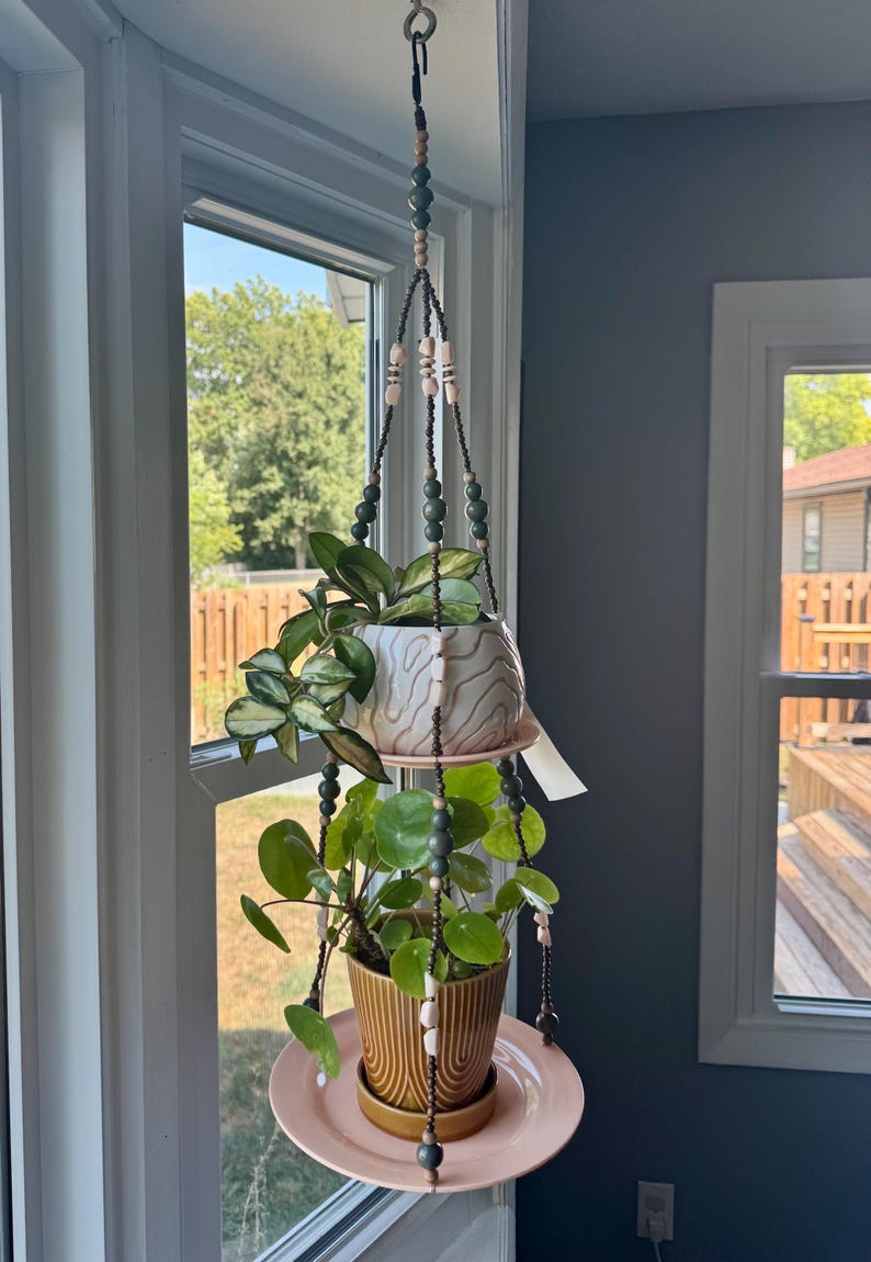 May include: A hanging plant holder with two ceramic pots and a pink plate. The holder is suspended from a hook and adorned with beads. The pots contain green plants, creating a decorative indoor display.