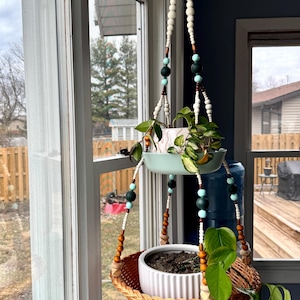 May include: Two hanging plant pots with a woven wicker basket and a light blue ceramic pot. The pots are suspended from a macrame-style beaded cord with white, brown, and green beads.