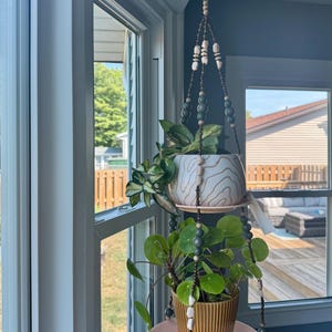 May include: A hanging macrame plant holder with two ceramic pots, one with a white and brown swirl design and the other with a ribbed, mustard-coloured finish. The holder features wooden beads and holds two plants with green foliage. The pots sit on pink saucers.
