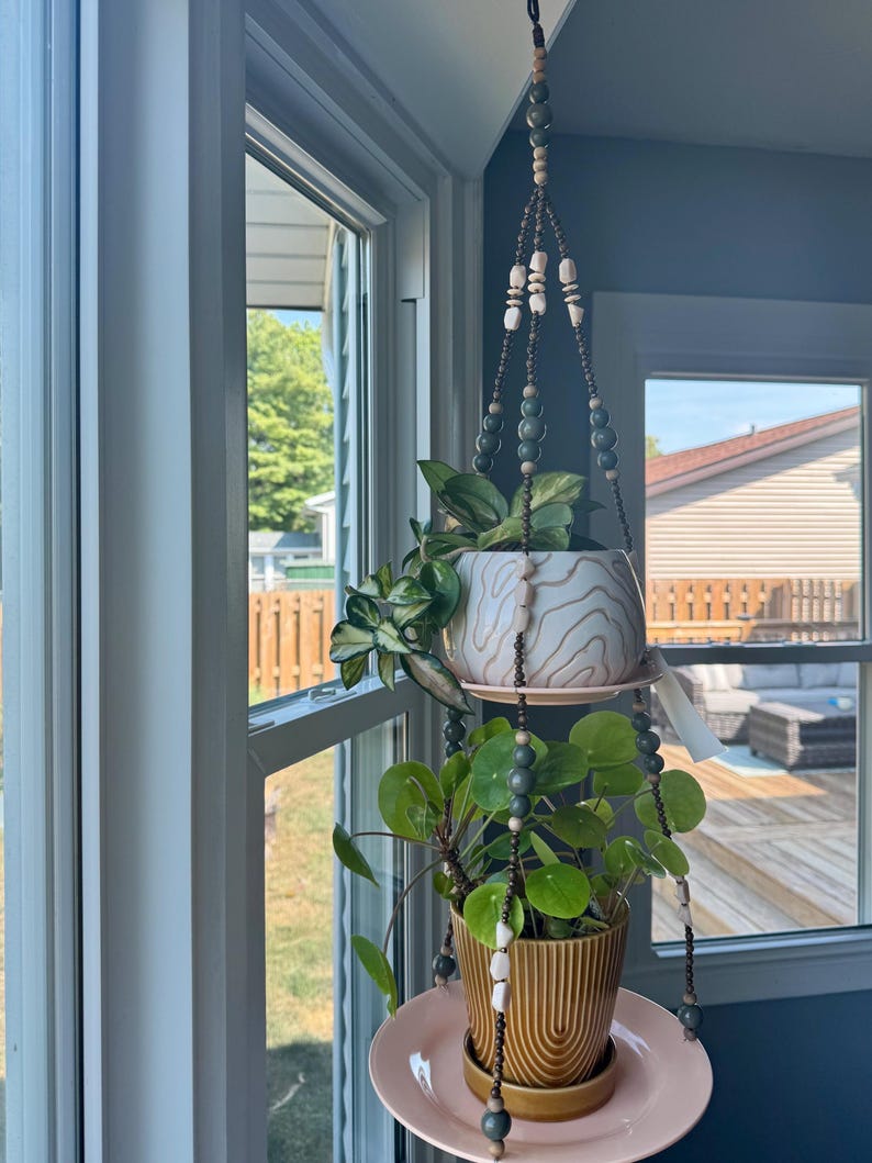 May include: A hanging macrame plant holder with two ceramic pots, one with a white and brown swirl design and the other with a ribbed, mustard-colored finish. The holder features wooden beads and holds two plants with green foliage. The pots sit on pink saucers.