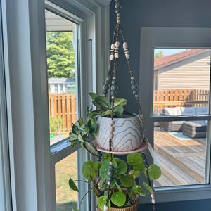 May include: A hanging plant holder with two levels, suspended from a ceiling hook. The holder features a beaded design with two pink plates supporting ceramic pots with plants. The top pot is white with a wood grain pattern.