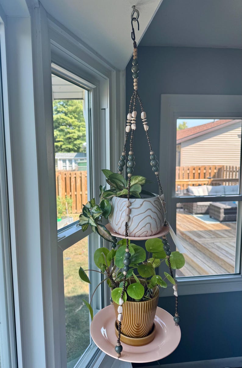 May include: A hanging plant holder with two levels, suspended from a ceiling hook. The holder features a beaded design with two pink plates supporting ceramic pots with plants. The top pot is white with a wood grain pattern.