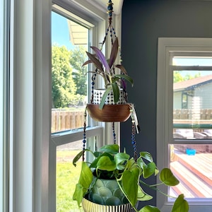 May include: A hanging plant holder with two levels, suspended from a ceiling hook. The top level holds a plant in a black and white patterned pot, and the bottom level has a green plant in a gold-colored bowl. The holder is decorated with blue and brown beads.