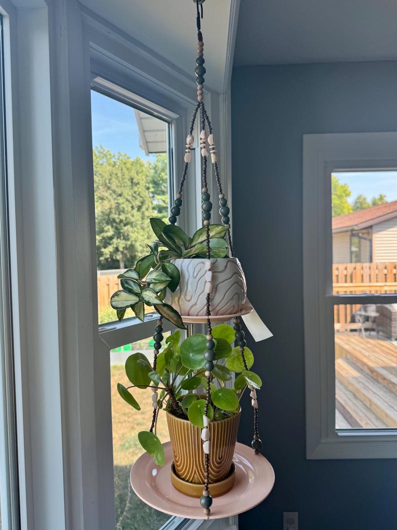 May include: A hanging plant holder with two ceramic pots, one white and one mustard yellow, suspended by beaded cords. The pots hold green plants and sit on pink saucers. The holder is suspended from a hook near a window.
