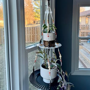 Può includere: Un portapiante sospeso in macramè con due vasi in ceramica bianchi, ciascuno con una pianta verde. Il supporto presenta perline decorative nei toni del bianco, rosa e legno. I vasi poggiano su vassoi di metallo nero, vicino a una finestra.
