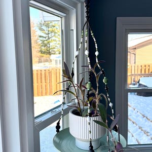 以下が含まれることがあります： 白いセラミックの鉢と薄緑色の受け皿が付いた、吊り下げ式の植物ホルダー。ホルダーは、黒、白、緑のビーズチェーンで天井から吊り下げられています。植物は紫と緑の葉を持っています。