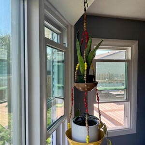 May include: A hanging plant holder with two tiers, suspended from a ceiling hook. The top tier is a brown bowl, and the bottom tier is a yellow bowl. Both hold potted plants. The hanging cords are adorned with red, yellow, and wooden beads.