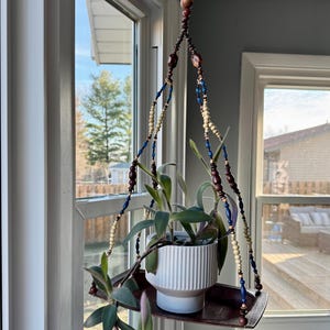 May include: A hanging plant holder with a white ceramic pot and a dark wooden tray. The hanger is made of beaded strands in shades of brown, blue, and cream. The plant has green and purple leaves, and the scene is set near a window.
