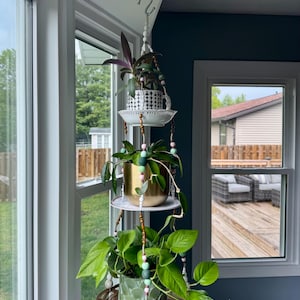 May include: A three-tiered hanging planter with various potted plants. The top level features a black and white patterned teacup. The middle level has a gold pot, and the bottom level has a green textured pot. The hanging structure is adorned with beads.