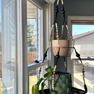 May include: A two-tiered plant hanger made of black and natural wood beads with a black metal frame. The hanger is holding two potted plants, one with green leaves and one with a green and white patterned pot.