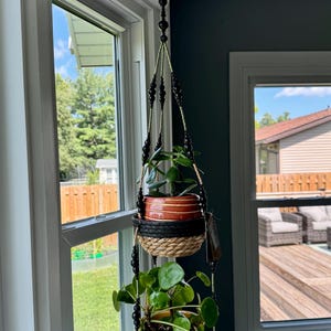 May include: A hanging plant holder with three tiers, suspended from a ceiling hook. The top tier holds a terracotta pot, the middle tier has a woven basket, and the bottom tier features a light green, scalloped planter. The holder is adorned with black beads.