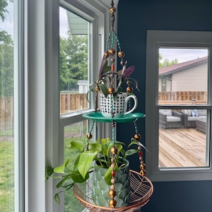 May include: A hanging plant holder with two tiers. The top tier holds a teacup and saucer with a black and white pattern, and the bottom tier has a green textured pot in a copper wire basket. The holder is suspended by a beaded chain.
