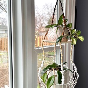 May include: A hanging plant holder with two levels, suspended from a black cord. The top level holds a white ceramic pot with a decorative design, while the bottom level features a white wire basket. Both levels display green plants.