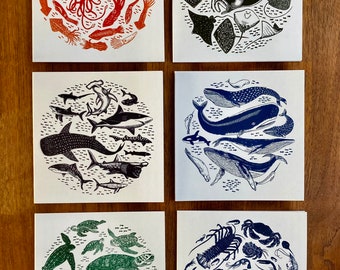 May include: Set of five illustrated cards featuring different sea creatures in a circular design. The cards are printed on white paper with a subtle texture. The designs include a red squid, a black manta ray, a blue whale, a green sea turtle, and a blue lobster.