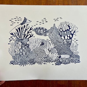 Puede incluir: Una impresión en linóleo azul y blanco de una escena de arrecife de coral con una tortuga marina, una raya y varios peces. La impresión está en papel blanco y tiene una superficie texturizada.