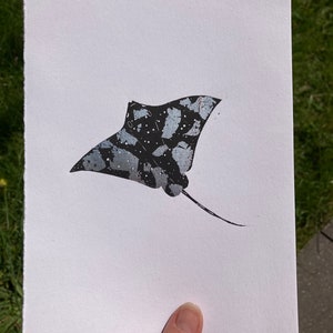 May include: A black and white print of a manta ray with a silver glitter finish. The manta ray is swimming with its wings spread out.