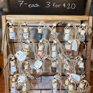 May include: A wooden display rack holds multiple car air fresheners. Each small glass bottle is suspended by a natural fibre cord. The sign above reads "CAR DIFFUSER". The bottles contain various coloured liquids and decorative elements.