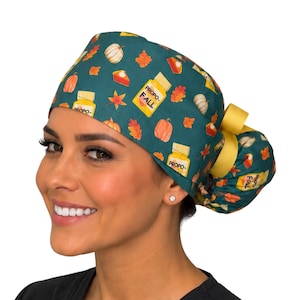 May include: A dark teal scrub cap featuring a repeating pattern of orange pumpkins, red and orange autumn leaves, slices of pumpkin pie, and yellow medicine bottles labeled PROPO-FALL. The cap includes a yellow ribbon tie at the back.