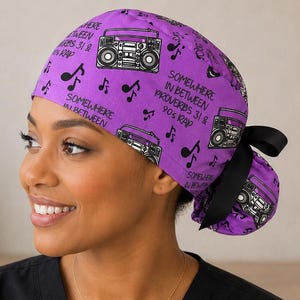 May include: A purple scrub cap featuring a repeating pattern of black boomboxes and musical notes. The text "SOMEWHERE IN BETWEEN PROVERBS 31 & 90s RAP" is printed in black. The cap includes a black ribbon tie at the back to secure the fit.