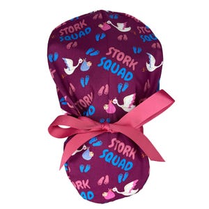 Könnte beinhalten: Eine lila OP-Mütze mit einer rosa Schleife. Die Mütze hat ein sich wiederholendes Muster aus Störchen, die Babys tragen, und Fußabdrücken. Der Text "Stork Squad" wird auf der Mütze wiederholt.