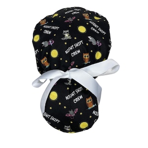 Puede incluir: Un gorro de quirófano negro con una cinta blanca. El gorro tiene un patrón repetido de búhos, mapaches, murciélagos, lunas y estrellas. El texto "Night Shift Crew" se repite en todo el patrón.