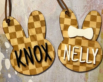 May include: Two wooden bunny-shaped ornaments with a checkered pattern. One ornament has the name "KNOX" in black lettering and the other has the name "NELLY" in white lettering with a white bow.