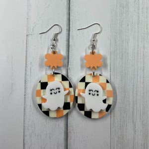 K&ouml;nnte beinhalten: Ein Paar Halloween-Ohrh&auml;nger. Jeder Ohrring hat einen wei&szlig;en Geisteranh&auml;nger auf einem schwarz-orange-cremefarbenen karierten Hintergrund. Die Ohrringe sind durch orangefarbene bl&uuml;tenf&ouml;rmige Akzente und silberne Haken verbunden.