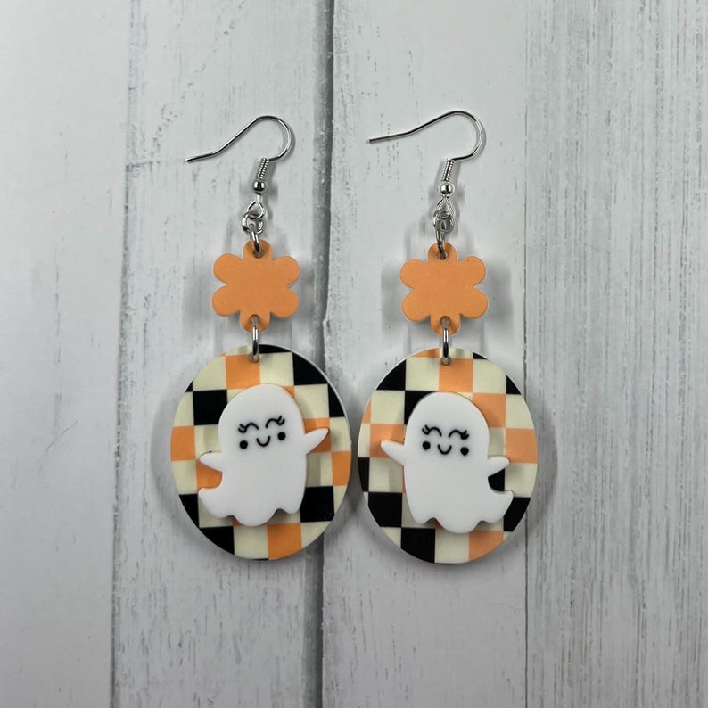 K&ouml;nnte beinhalten: Ein Paar Halloween-Ohrh&auml;nger. Jeder Ohrring hat einen wei&szlig;en Geisteranh&auml;nger auf einem schwarz-orange-cremefarbenen karierten Hintergrund. Die Ohrringe sind durch orangefarbene bl&uuml;tenf&ouml;rmige Akzente und silberne Haken verbunden.