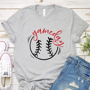 May include: A gray t-shirt with the text "gameday" in red cursive writing and a black baseball graphic. The shirt is laying on a white wooden surface with a pair of white sneakers and blue denim shorts.