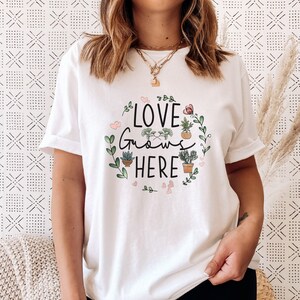 May include: White t-shirt with a green and pink graphic that says "Love Grows Here" with potted plants and hearts.