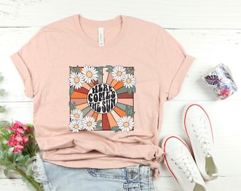 May include: A heather peach t-shirt with a retro graphic design featuring a sun with rays and the text "Here Comes The Sun" surrounded by white daisies.