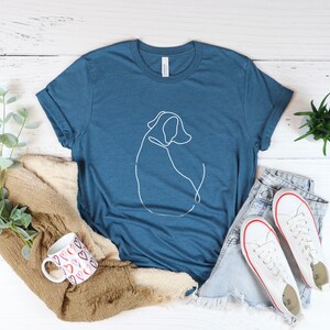 May include: A heather deep teal t-shirt with a white line drawing of a dog on the front. The shirt is laying on a white wooden surface with a pair of white sneakers, a mug, and a plant.