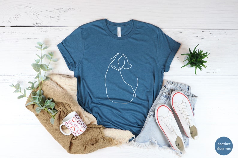 May include: A heather deep teal t-shirt with a white line drawing of a dog on the front. The shirt is laying on a white wooden surface with a pair of white sneakers, a mug, and a plant.