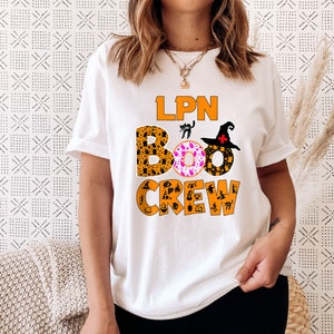 Pode incluir: Camiseta branca com o texto "LPN BOO CREW" em laranja e preto. O texto &eacute; cercado por &iacute;cones tem&aacute;ticos de Halloween, incluindo um chap&eacute;u de bruxa, um gato preto e uma ab&oacute;bora.