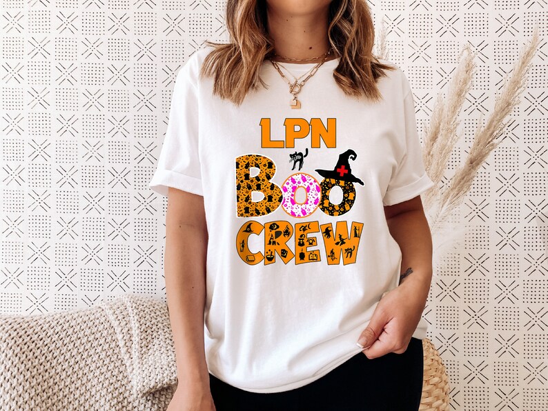 Pode incluir: Camiseta branca com o texto "LPN BOO CREW" em laranja e preto. O texto &eacute; cercado por &iacute;cones tem&aacute;ticos de Halloween, incluindo um chap&eacute;u de bruxa, um gato preto e uma ab&oacute;bora.