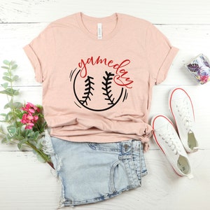May include: A heather peach t-shirt with a black and white baseball graphic and the text "gameday" in red.