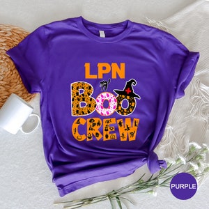 Pode incluir: Camiseta de manga curta roxa com o texto "LPN BOO CREW" em laranja e preto. O texto "BOO" &eacute; cercado por um padr&atilde;o rosa e laranja com &iacute;cones tem&aacute;ticos de Halloween.